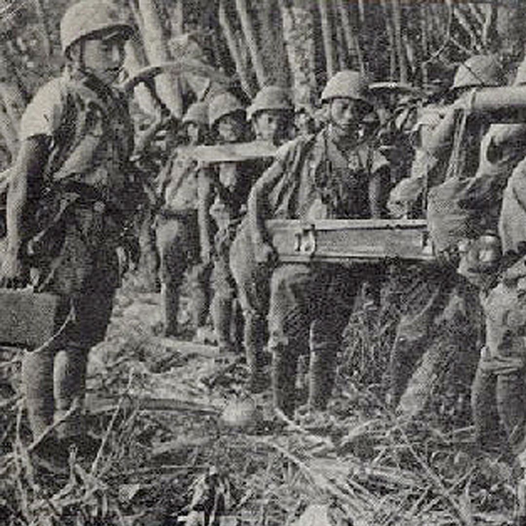 A grainy black and white photo of soldiers carrying large pieces of military equipment through the jungle.
