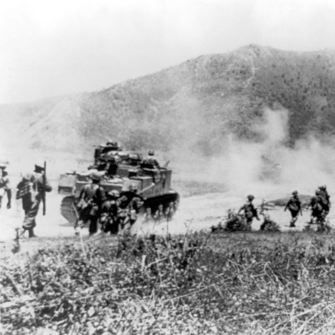 A black and white photograph of a tank advancing along a dusty road with Gurkha soldiers advancing along side.