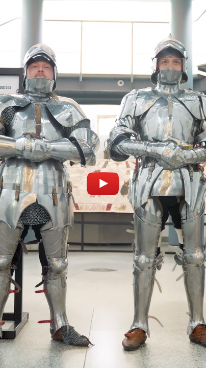 Clad in full suits of shiny armour, two people pose in front of a museum exhibit featuring a large window and decorative wall art; facial details are hidden by the armour