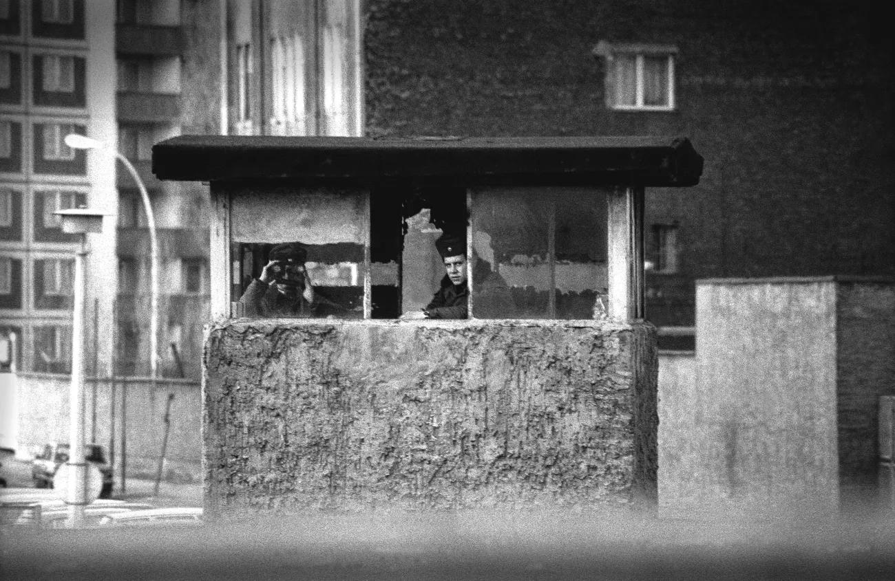 A black and white image showing two East German border guards sat in a concrete hut looking out of binoculars. They are surrounded by concrete plattenbau. 