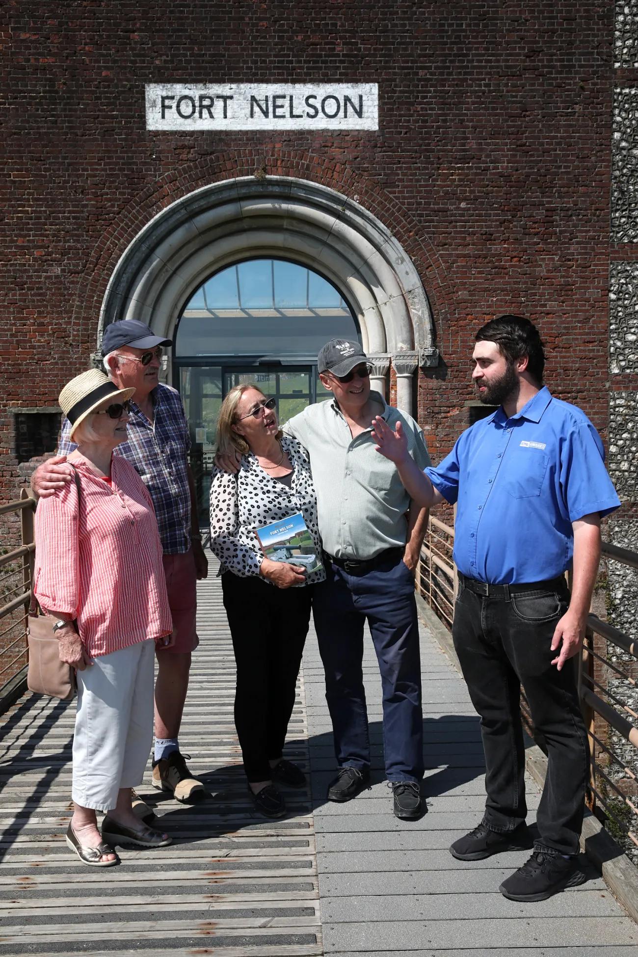 Group of individuals gathered on a bridge leading to Fort Nelson’s entrance, featuring a person with a book and another appearing to explain something