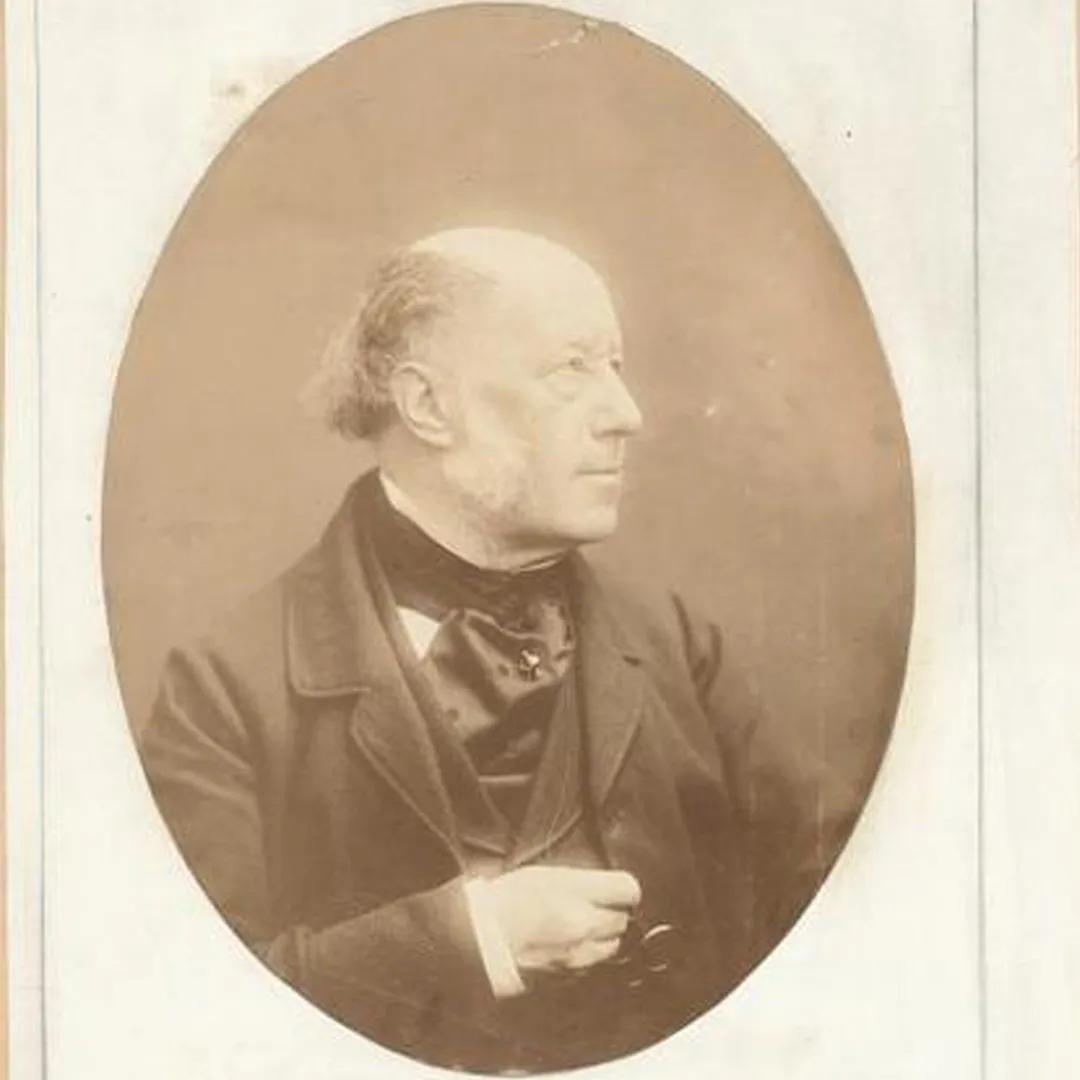 A sepia tone photograph of a balding man in a 19th century suit, glancing to one side and holding their pons nez.