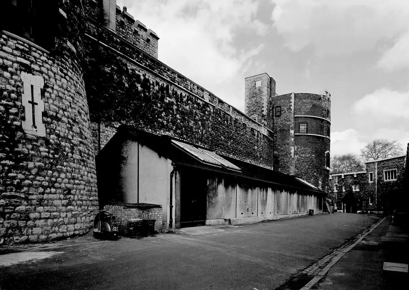 A black and white photograph of a long shed-like building set against a high stone wall