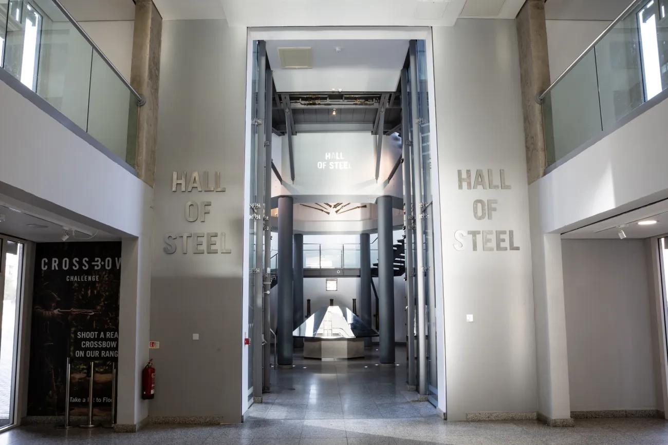 Two columns with silver lettering indicate the Hall of Steel. Inside, a large mirrored object sit in the centre of a hall, surrounded by cannons pinned upright to the wall. 