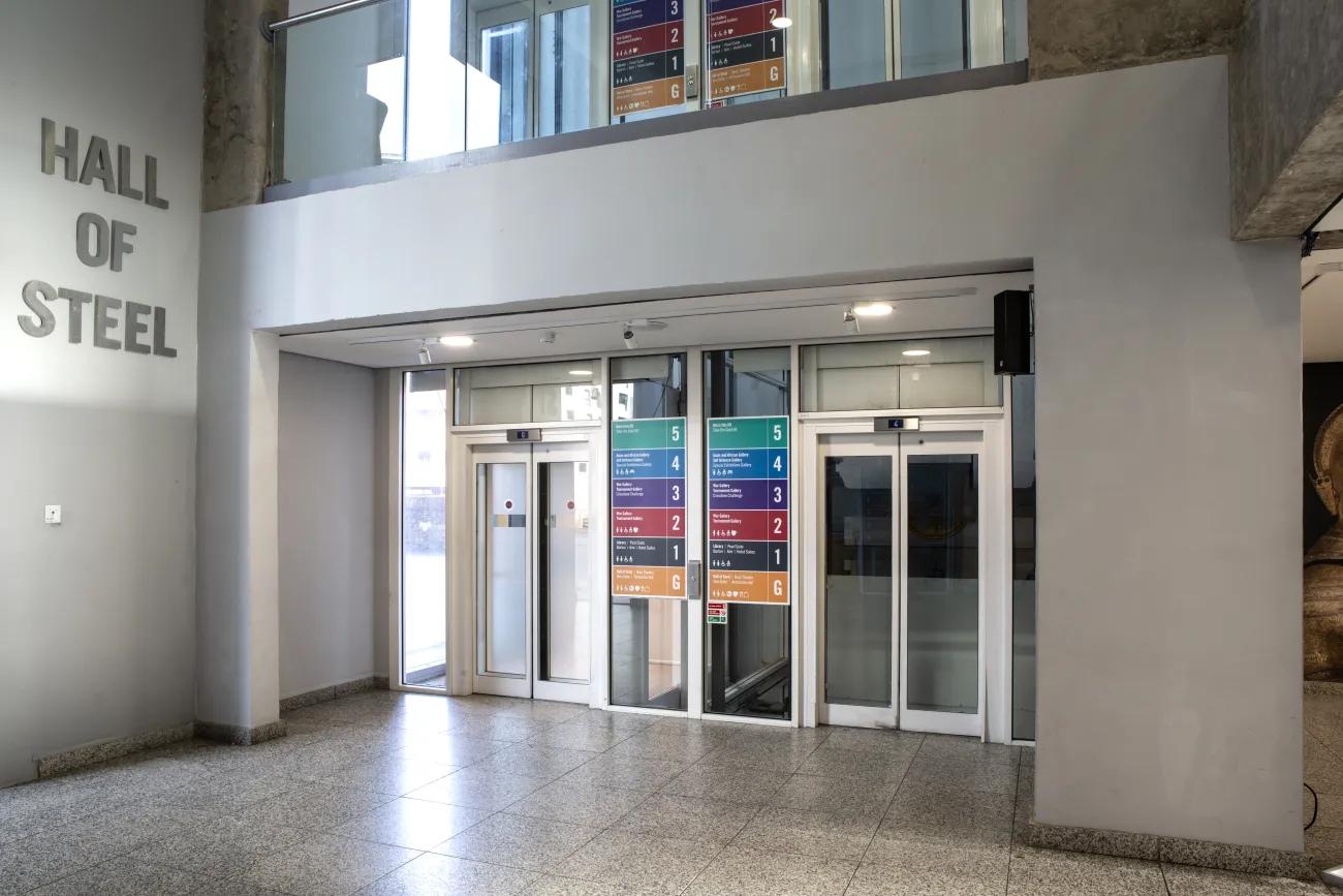 A grey floored hallway with two lifts in white facing. This set of lifts sits to the side of a large grey wall with metallic lettering reading 'Hall of Steel'.