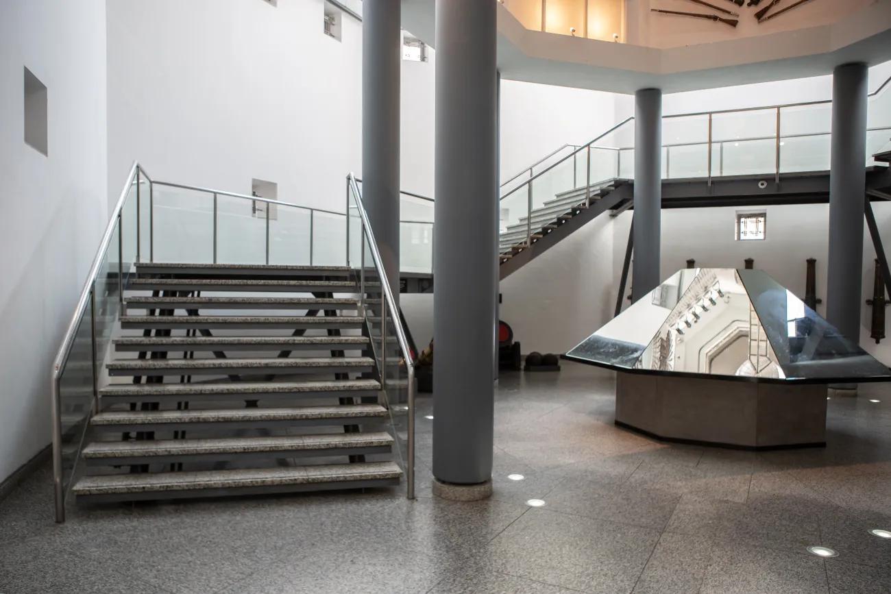 A flight of steel and glass stairs rise up and around the inside of a tower. In the centre of the ground floor of the tower sits a large mirror flanked by cannons pinned to the wall.