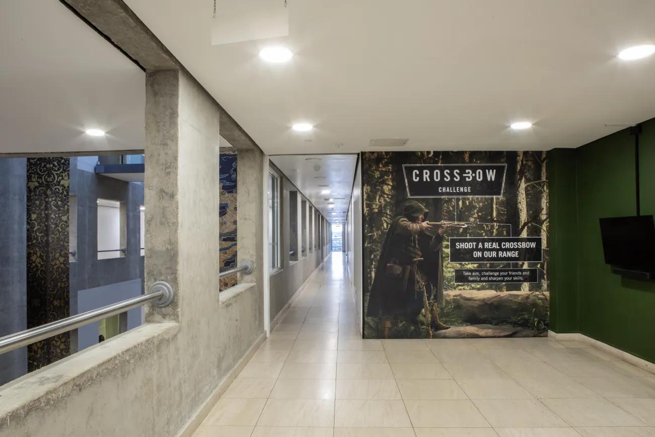 A grey corridor stretching beyond the camera, with a wall advertising a crossbow range beside it.