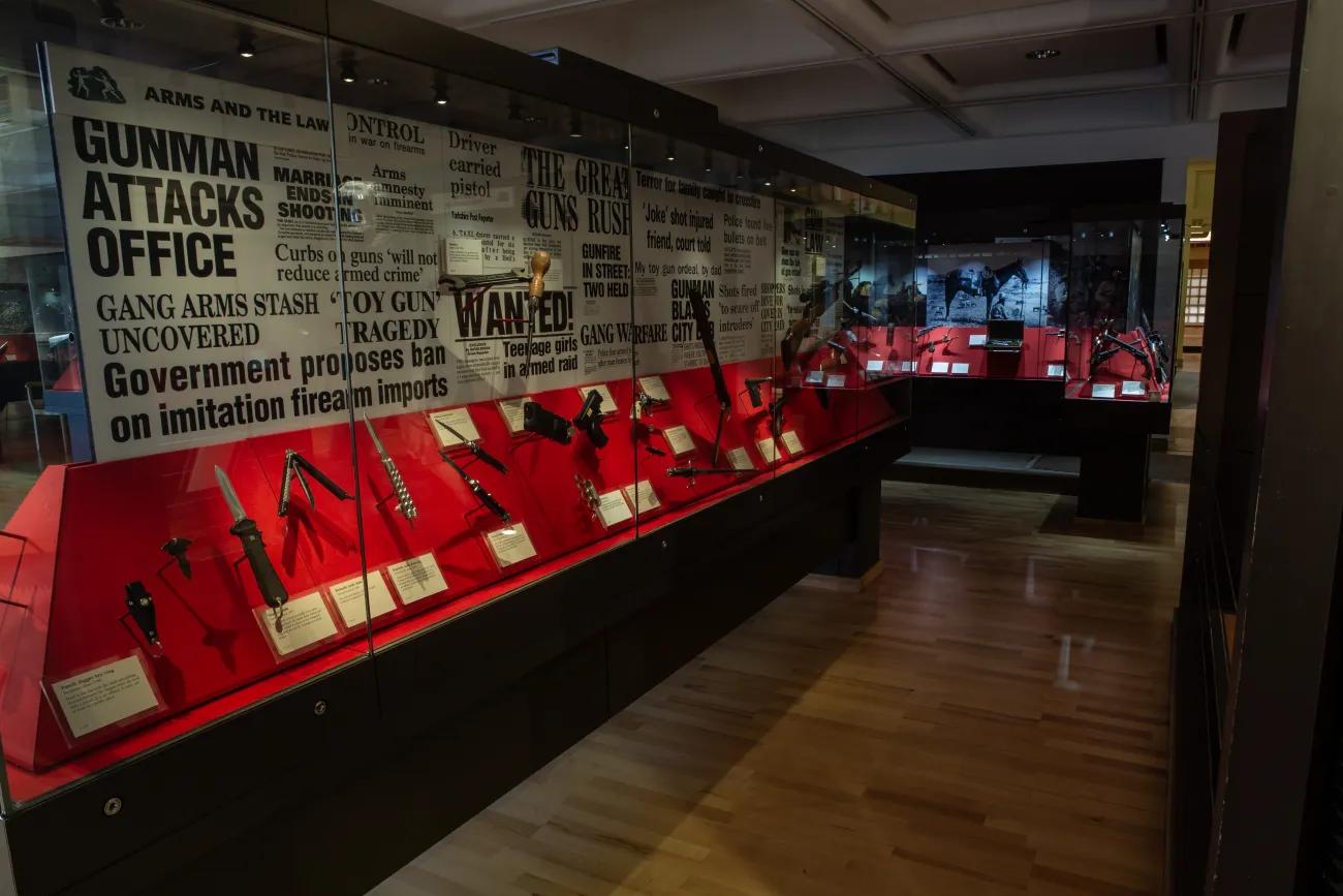 A dark room with a long, semi-lit cabinet containing a variety of knives and other compact weapons for self-defence