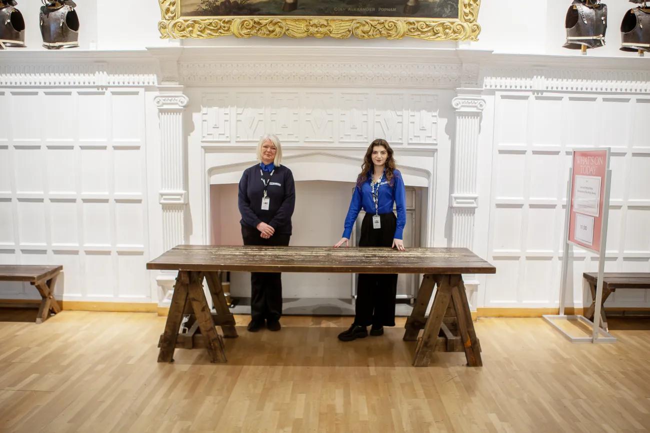 Two women in blue uniforms stand behind a wooden table in front of a fireplace
