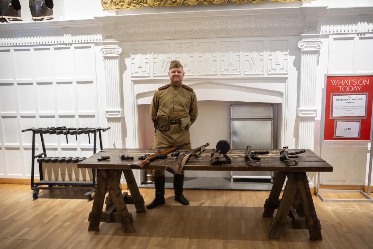 A visitor assistant in World War Two era Soviet battledress stands behind a table filled with Soviet weapons.