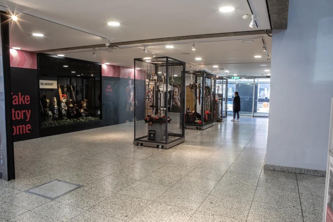 A wide hallway with cages filled with merchandise available to buy such as swords and helmets. At the far end of the hall, there is a set of closed glass doors. 