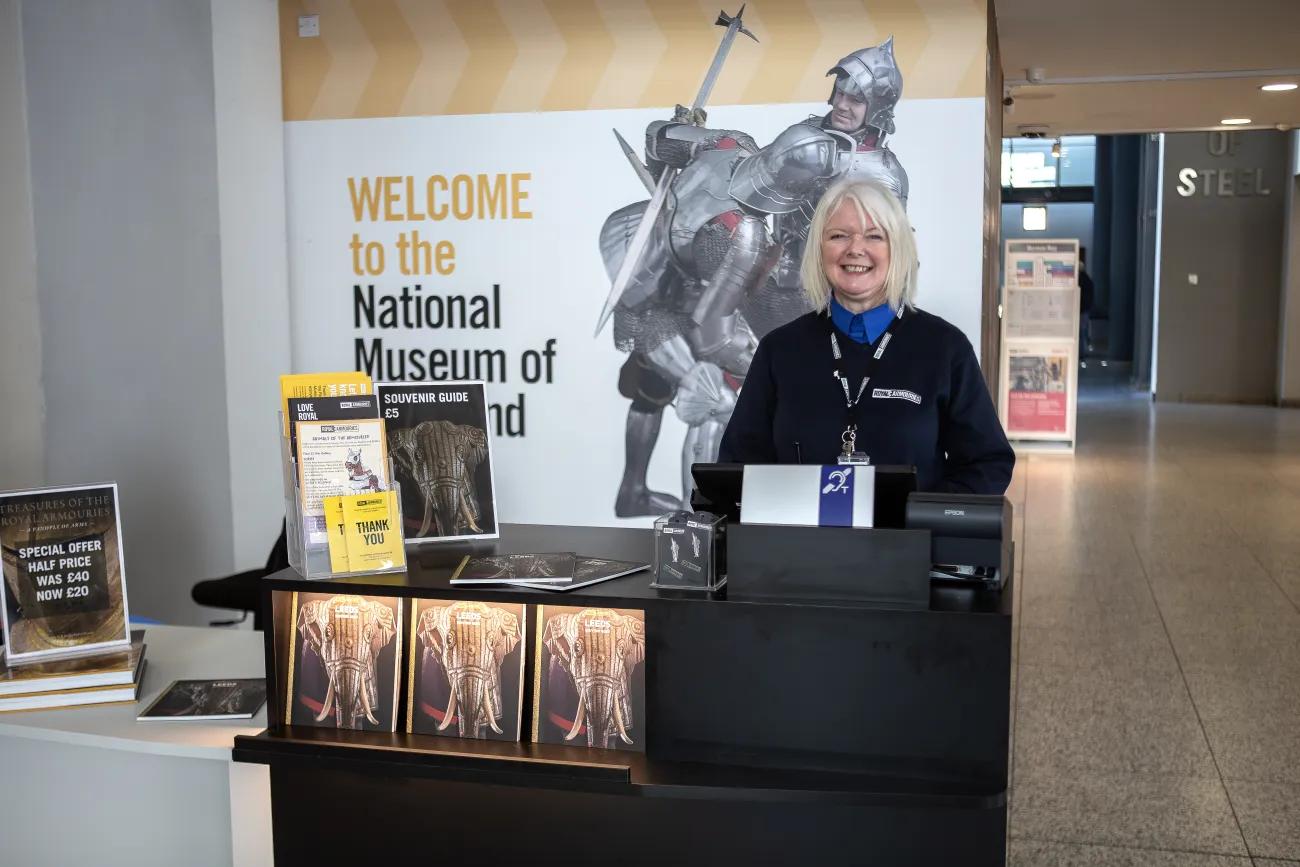 A woman in a blue uniform welcomes visitors behind a dark coloured desk. On the desk can be seen guide books to the museum. 