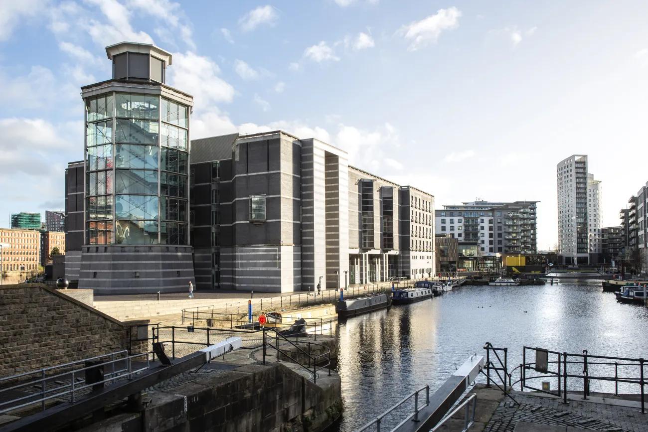 A colour photo of a large grey building beside a body of water. In front of and attached to the building is a large heptagonal glass tower with a staircase visible on the inside.  