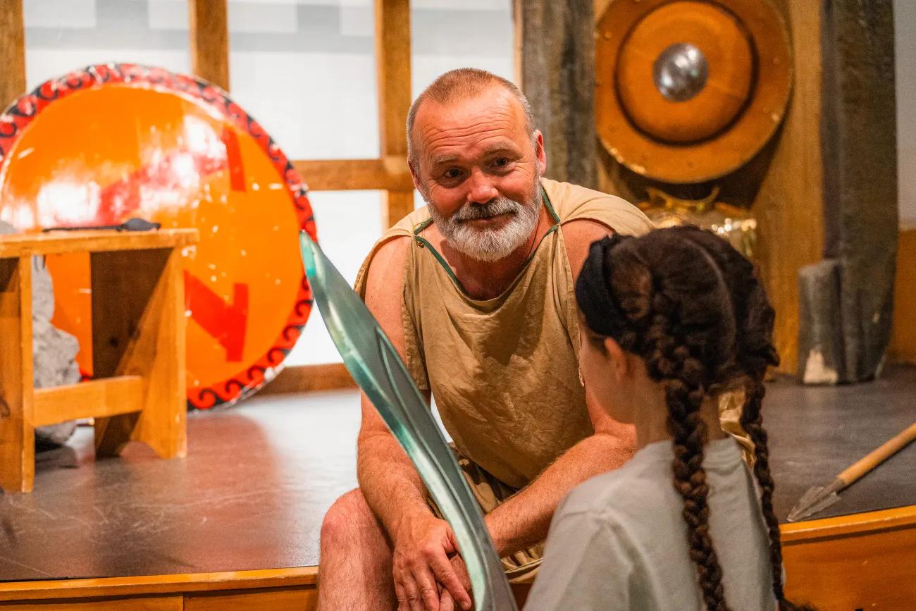 A man in a toga sits on a stage and instructs a young listener on how to wield a sword