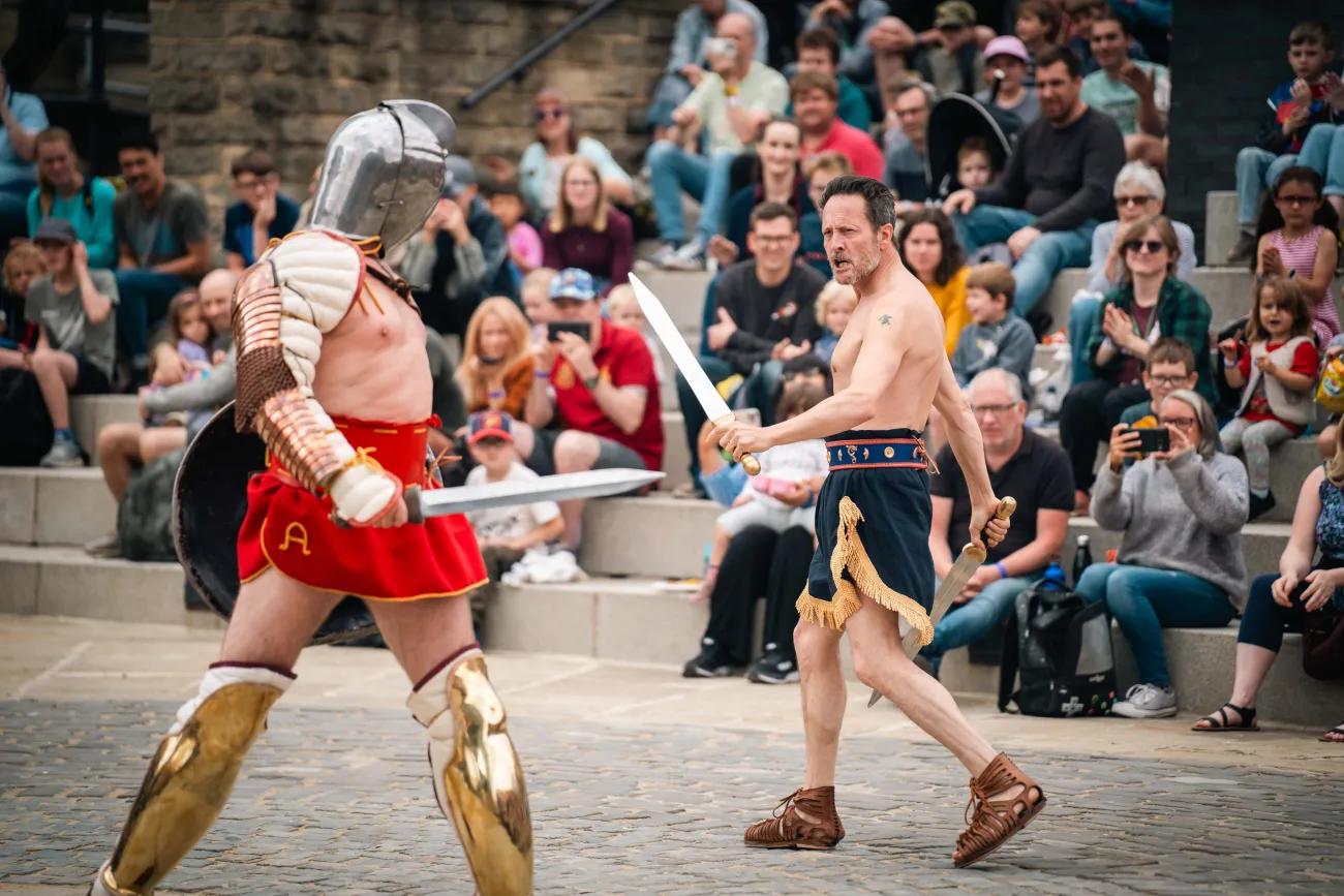 Two actors in Roman gladiatorial costume circle each other as a crowd looks on.