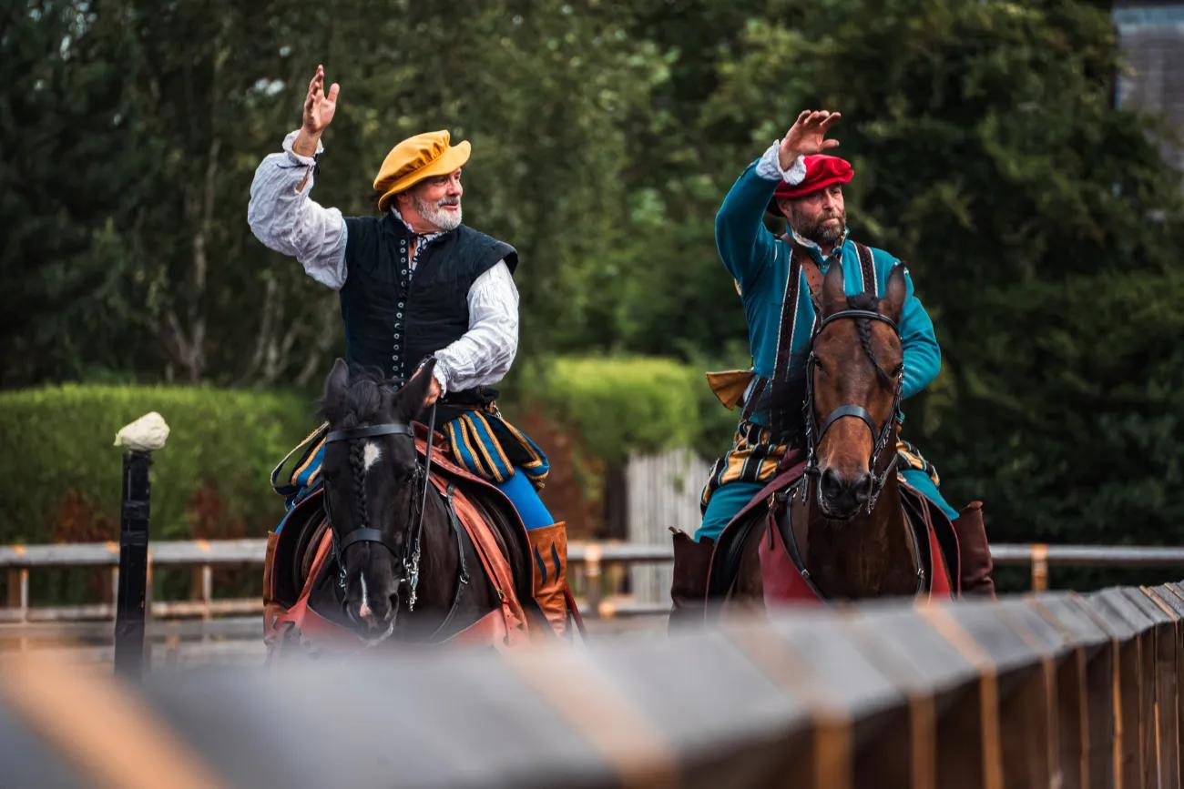 Two men on horseback and in Tudor era dress greet the crowd behind a wooden fence