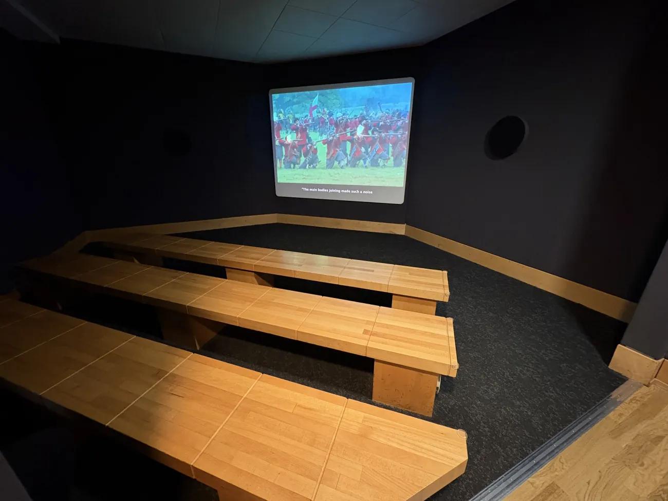 A dark room with plain wooden benches facing a screen showing a film about Marston Moor