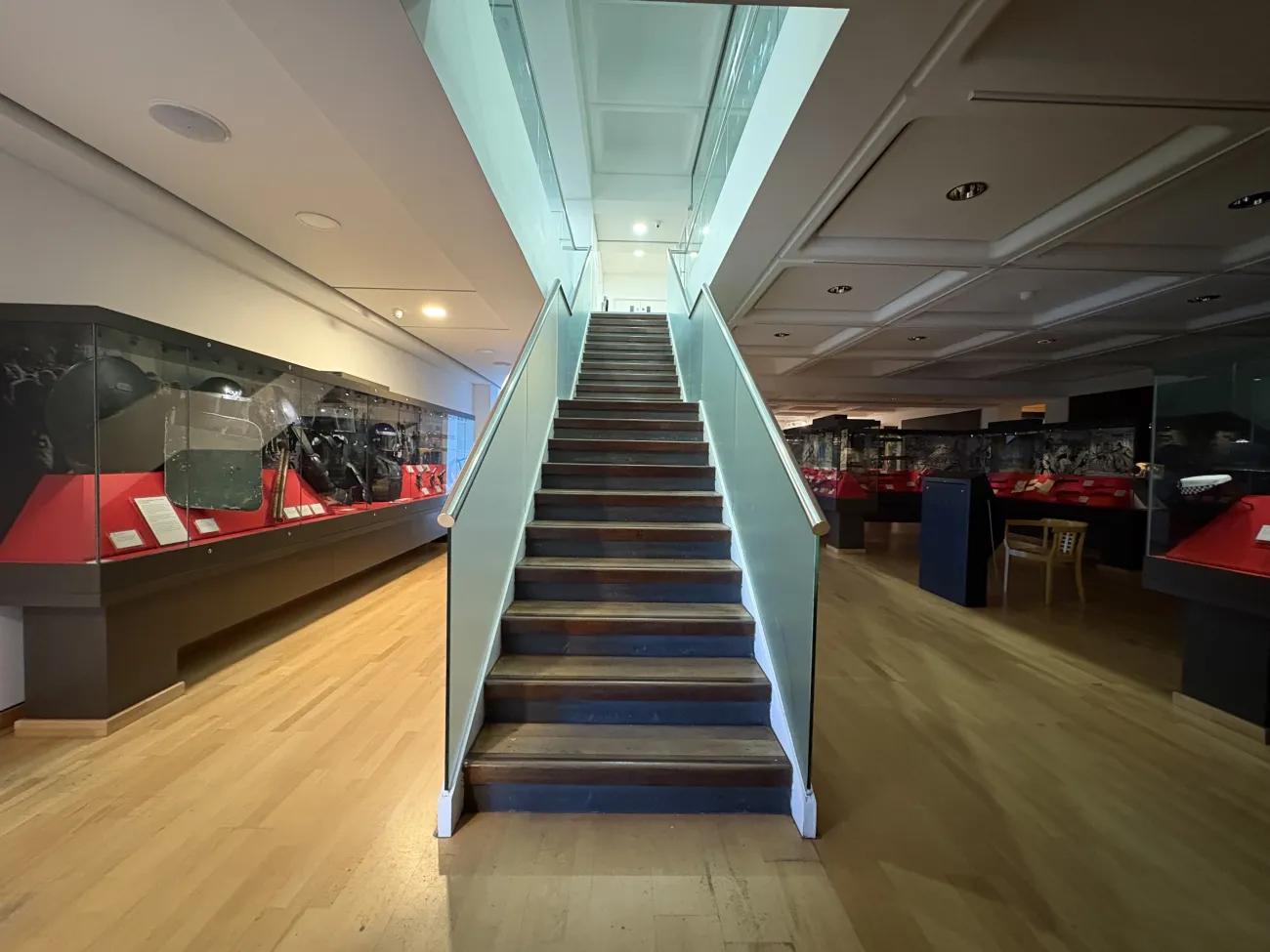 A darkened room with some sunlight coming in on the left hand side, lighting up a row of glass display cases. In the centre, a flight of stairs ascends away from the camera.