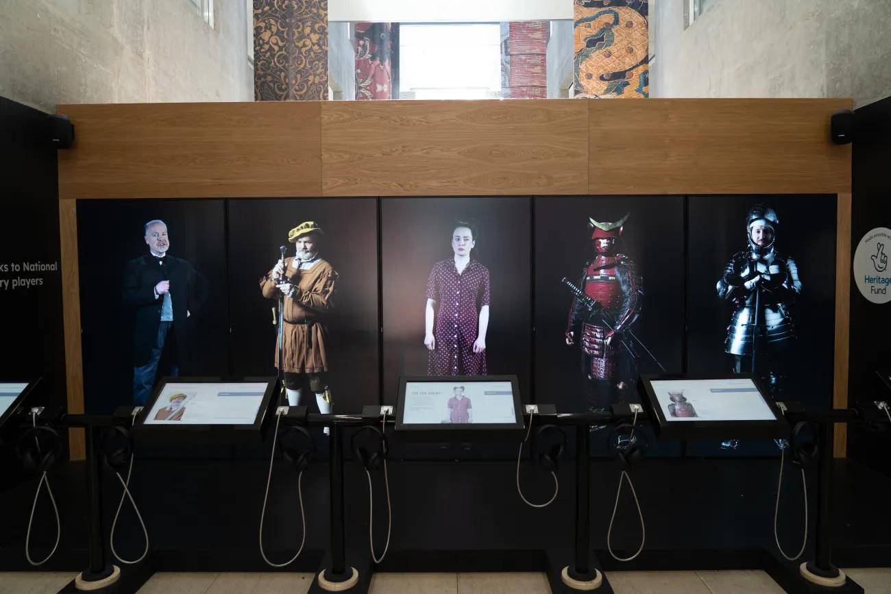 A wall of five human sized monitors displaying five characters from history, including a knight and a samurai in armour, a women in a purple dress from the 1940s, and a man in a Victorian suit.