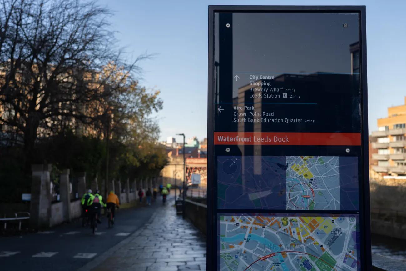 A dark blue and red sign with a map stood next to a pathway that runs by a river