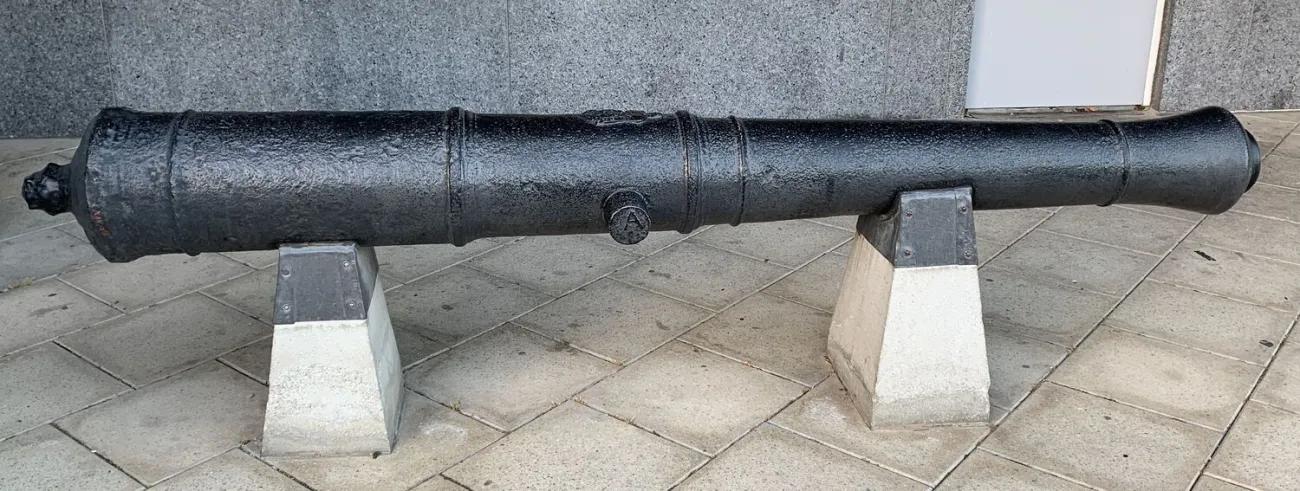 A weathered iron cannon with a rounded cascabel and central trunnions, mounted on two tapered concrete blocks and displayed on diagonally laid pavement tiles