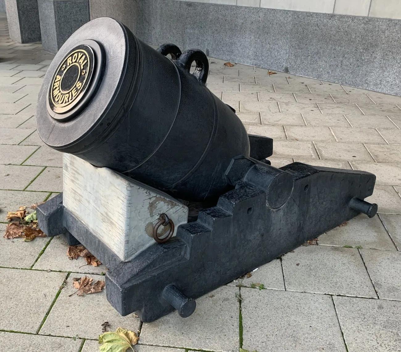 A large‑bore iron mortar with a decorative circular end plate marked “ROYAL ARMOURIES,” positioned on a black metal base and wooden block, displayed outdoors on patterned paving slabs