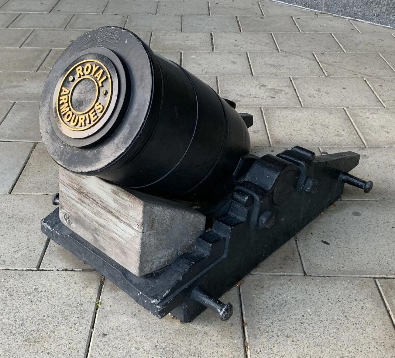 A large‑bore mortar painted black, featuring a decorative rear plate with raised lettering, positioned on a stepped metal carriage and timber block, displayed outdoors on evenly laid paving stones