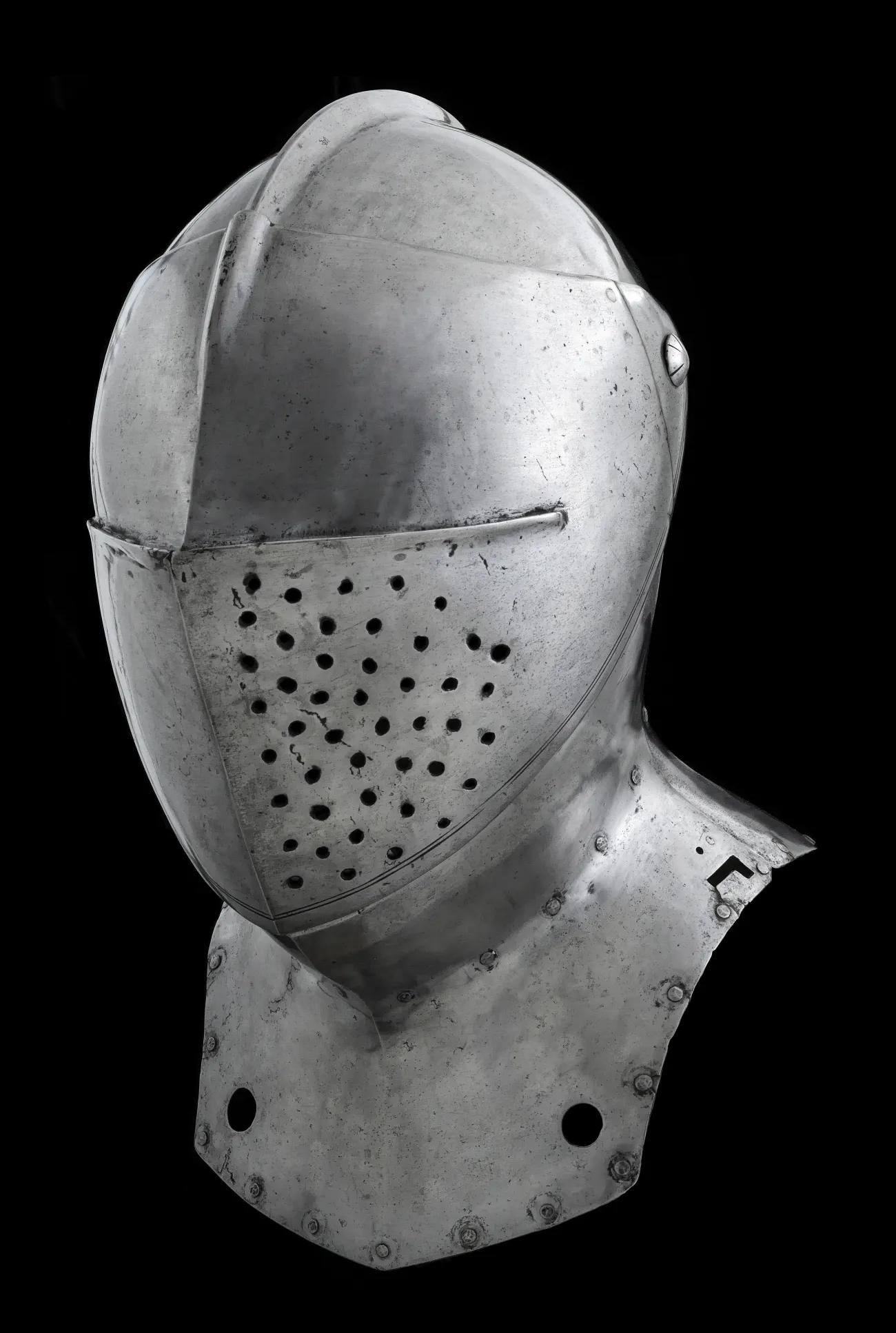A steel coloured metal helmet that encases the head. The helmet has pierced breathing holes but no sight holes of opening to allow the user to see. The background is entirely black.
