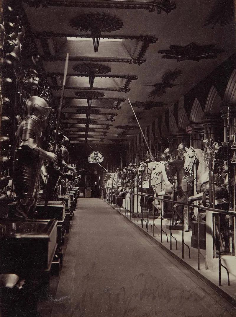 A sepia tone photograph of the interior of the New Horse Armoury, showing a line of armour mannequins on either side of a narrow aisle. 