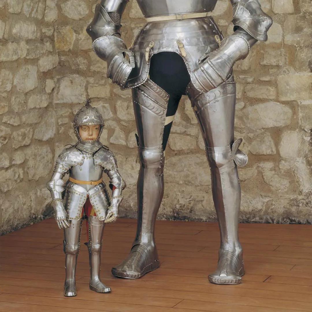 Two suits of armour stand next to each other. The first is very small, likely to fit a child. The second is very tall. Both are shining brightly.