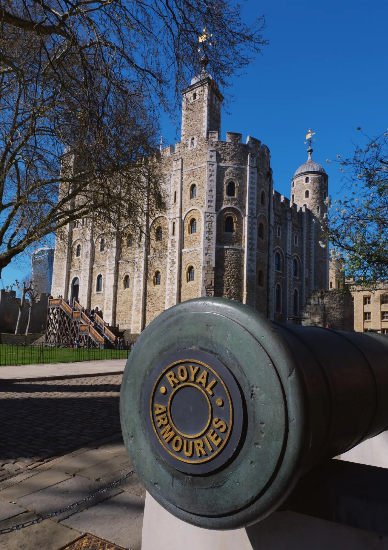 A colour photograph of the exterior of the White Tower at the Tower of London. There is a blackened cannon sat in the foreground with a plug saying Royal Armouries in gold lettering in the barrel.