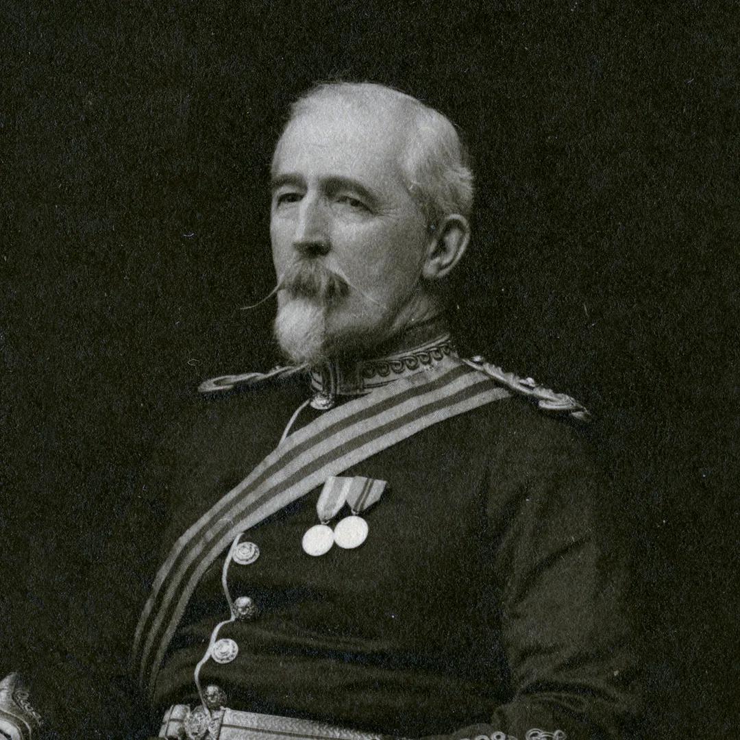 A black and white photo of a grey haired and bearded man in military uniform sat against a dark background