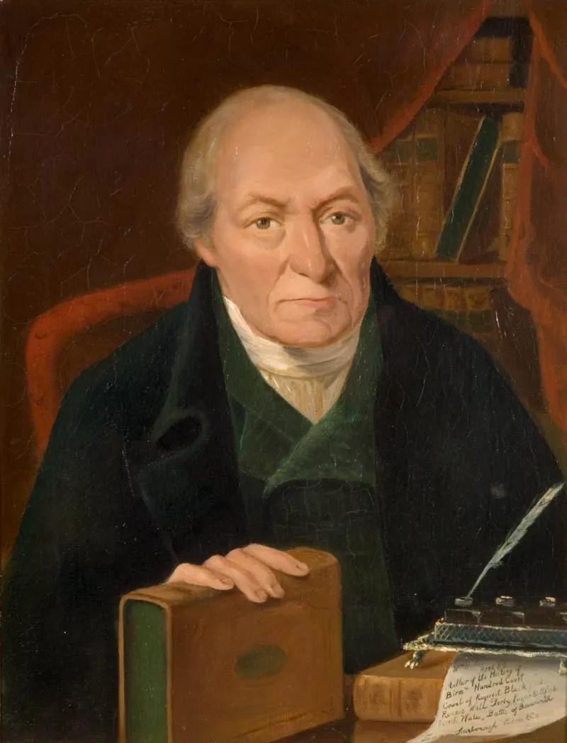 A colour portrait of a grim looking man in black frock coat and green waistcoat sat behind a desk with books, parchment and quill