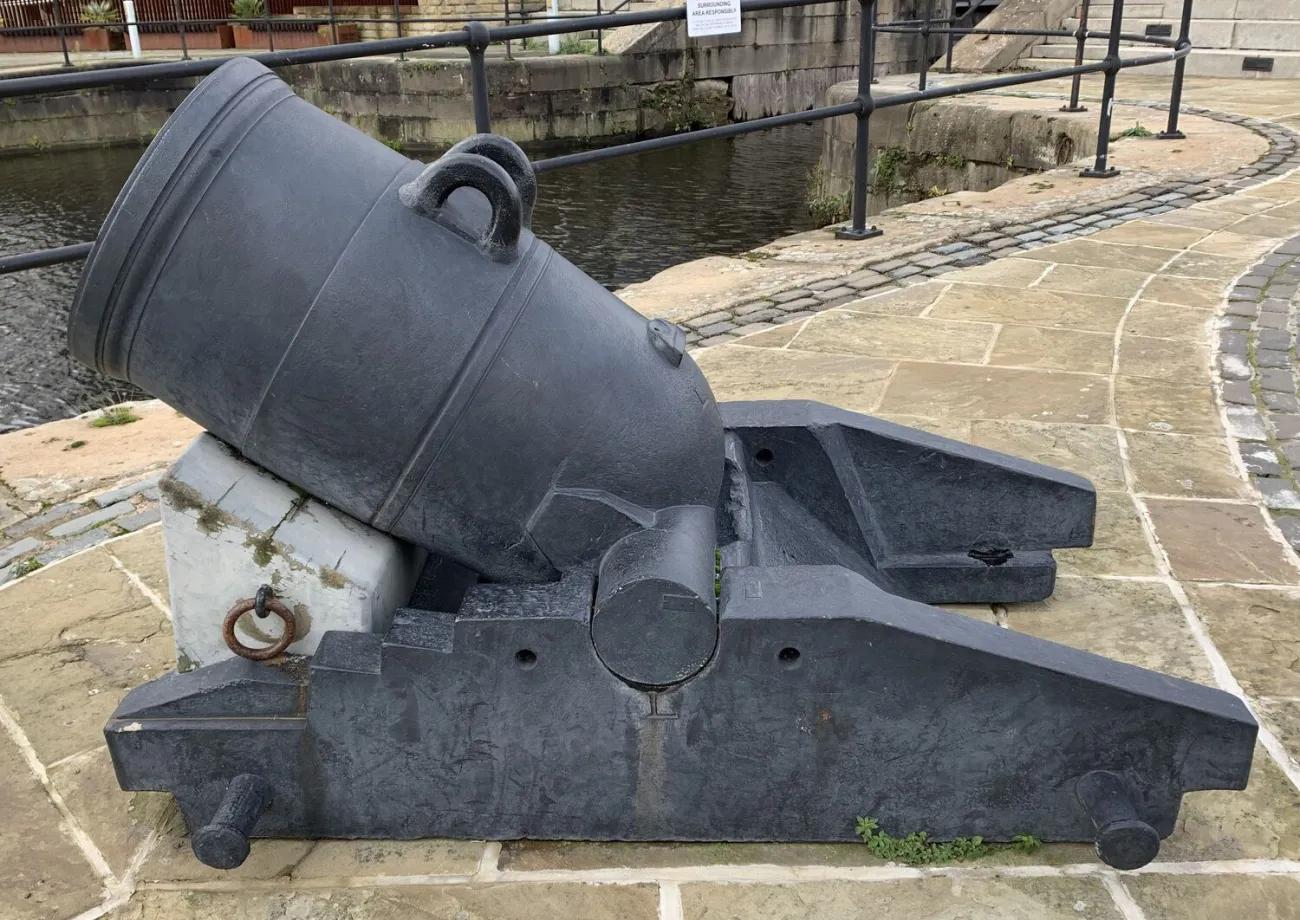 A historic artillery mortar with a wide bore and lifting rings, secured on a solid support frame, displayed outdoors near a canal edge with paving and railings surrounding it