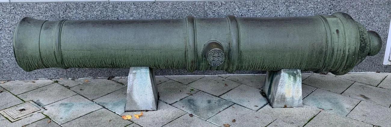 A long, weathered bronze cannon resting horizontally on two angular concrete supports, displayed on a paved area beside a granite wall, with a round decorative emblem on its side