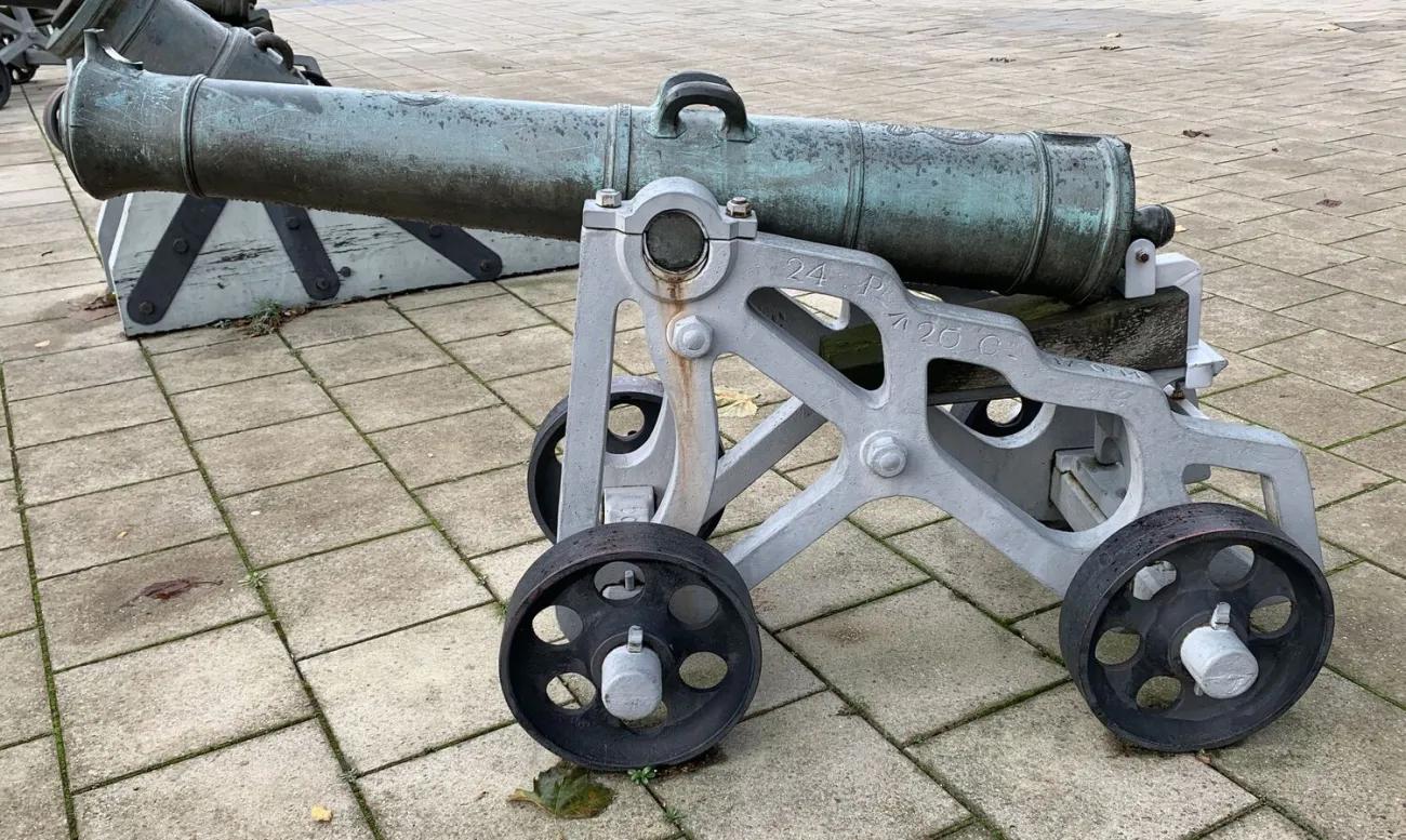 A short bronze‑green cannon with a weathered patina mounted on a pale grey metal carriage with four dark spoked wheels, standing on square paving stones with other artillery pieces behind