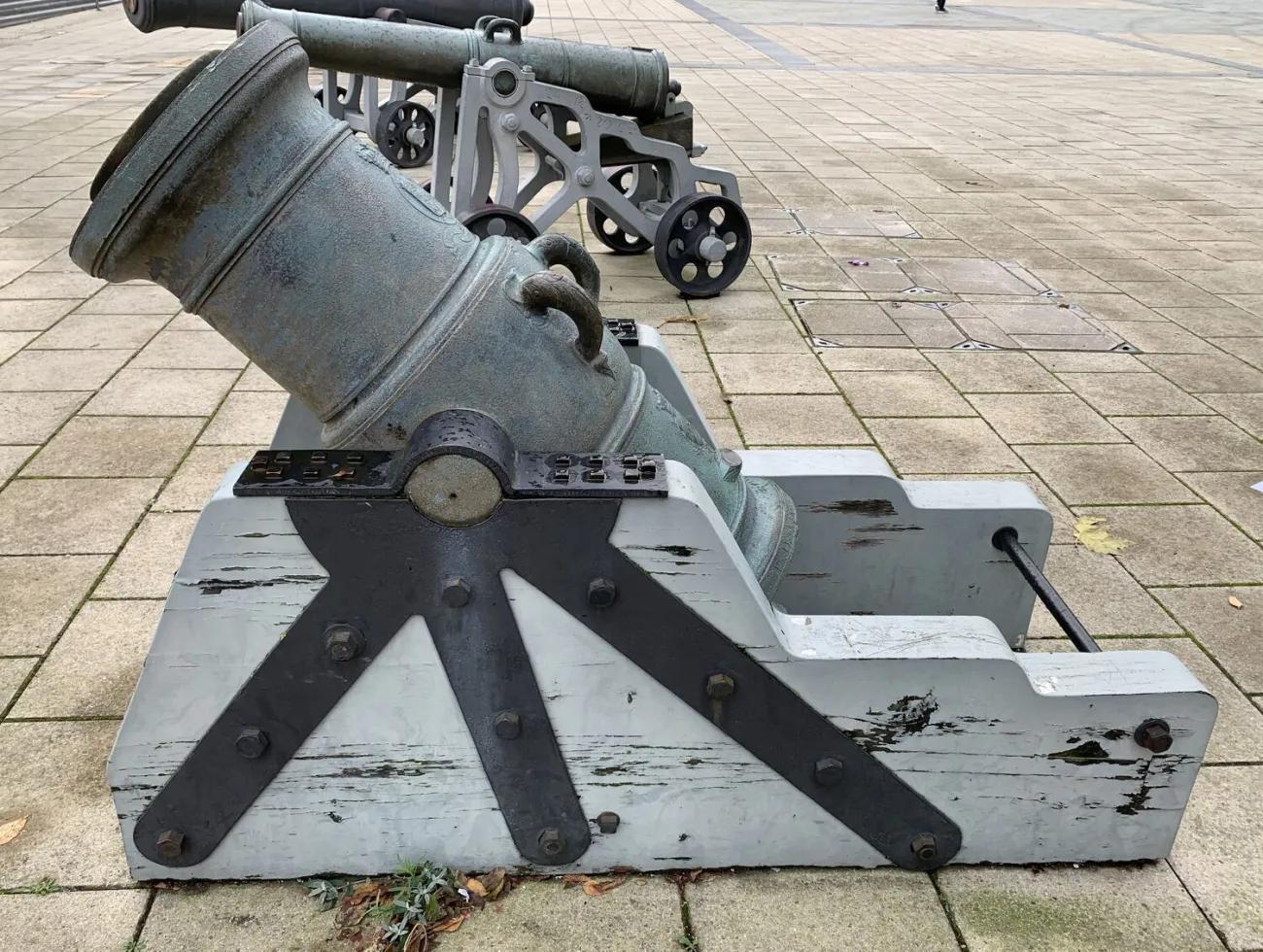 A heavy, aged mortar barrel set in a fixed angled wooden cradle painted pale grey and strengthened with dark metal supports, resting on a paved outdoor surface with additional cannon mounts in the background