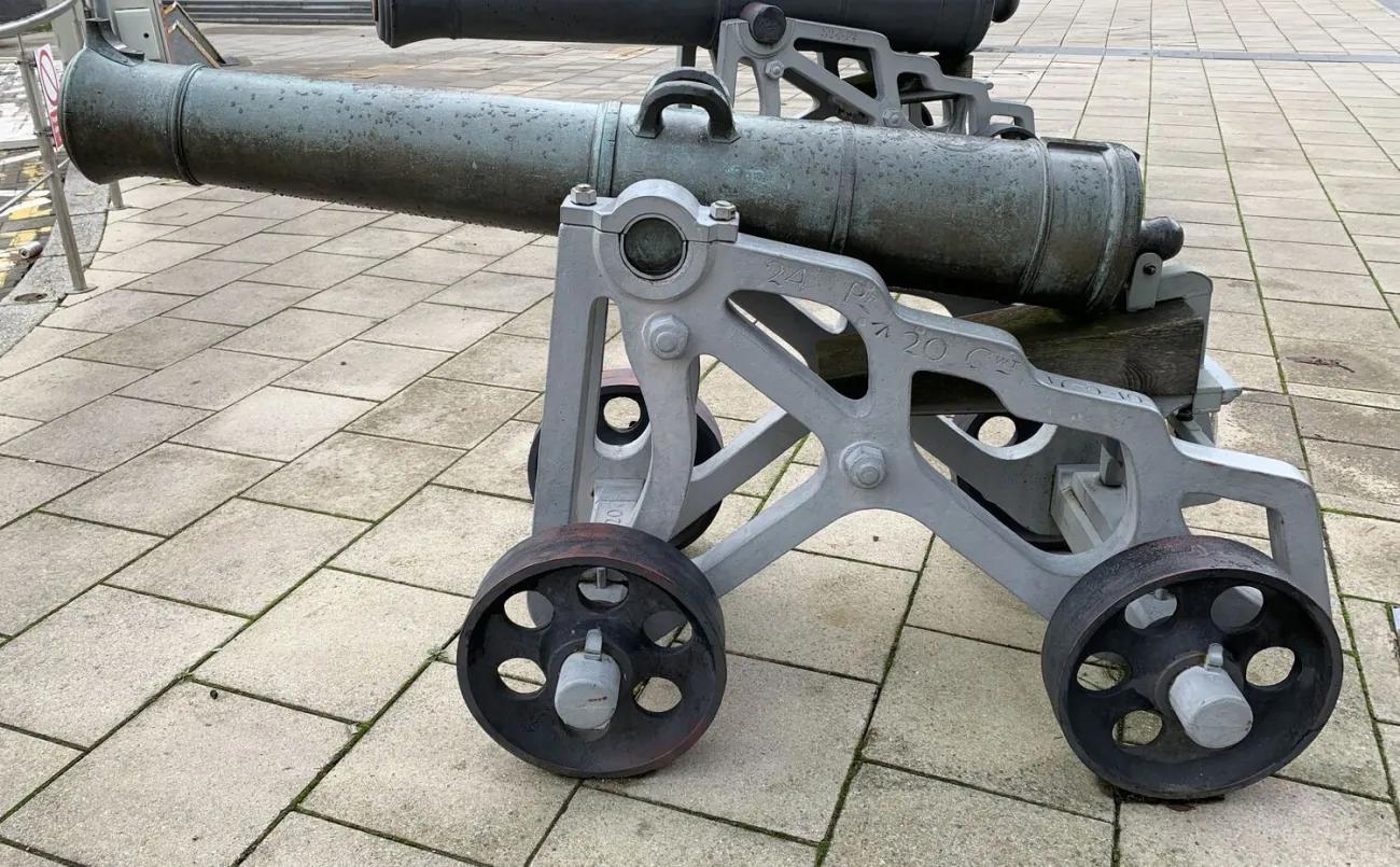 An aged bronze artillery barrel with visible green oxidation fixed to a modern grey wheeled carriage, placed on a broad paved area with another mounted cannon partially visible in the background