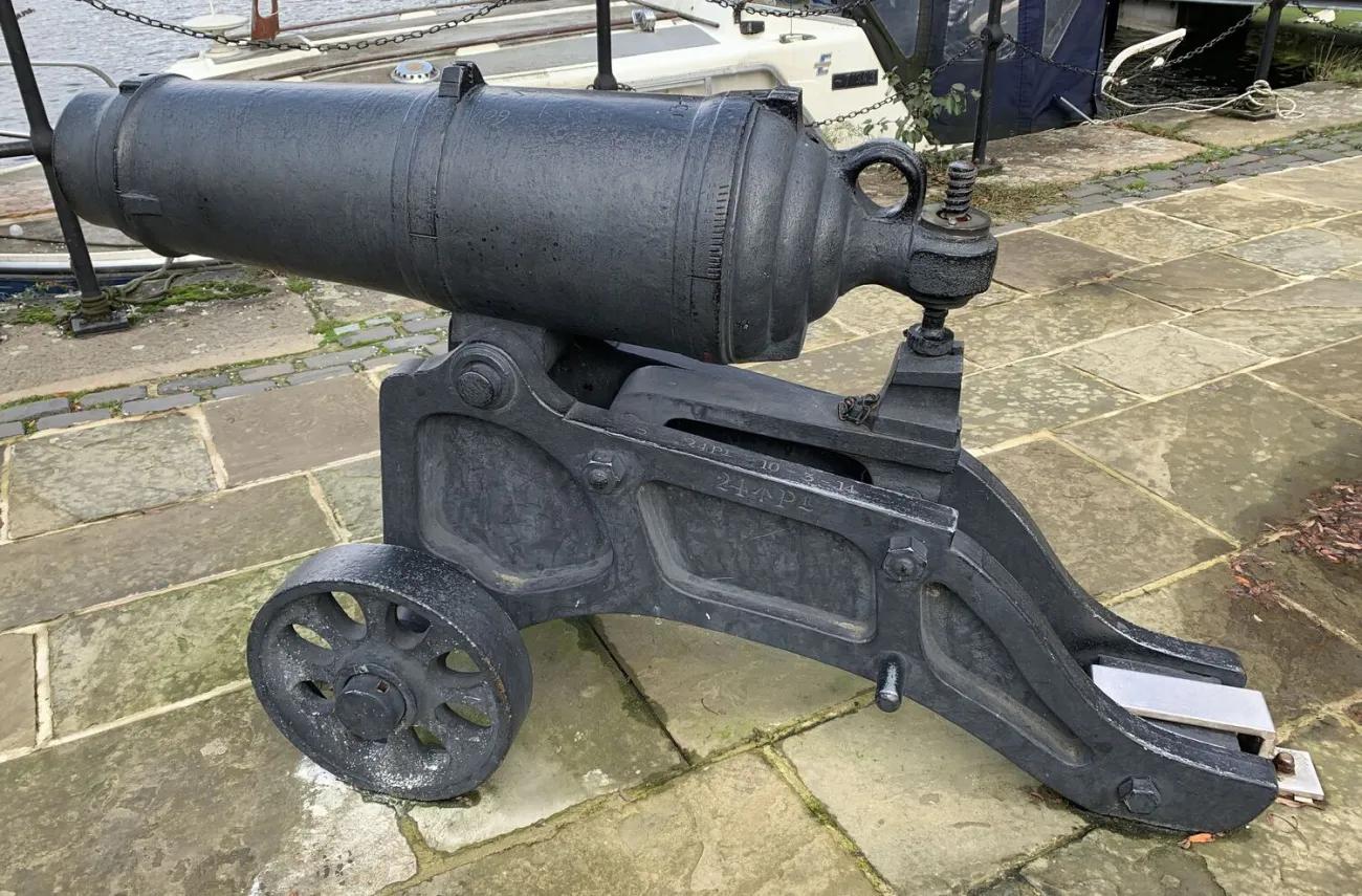 A compact, heavy artillery piece with a smooth dark barrel fixed to an arched metal carriage frame, supported by two round spoked wheels, positioned on stone slabs beside a waterside area