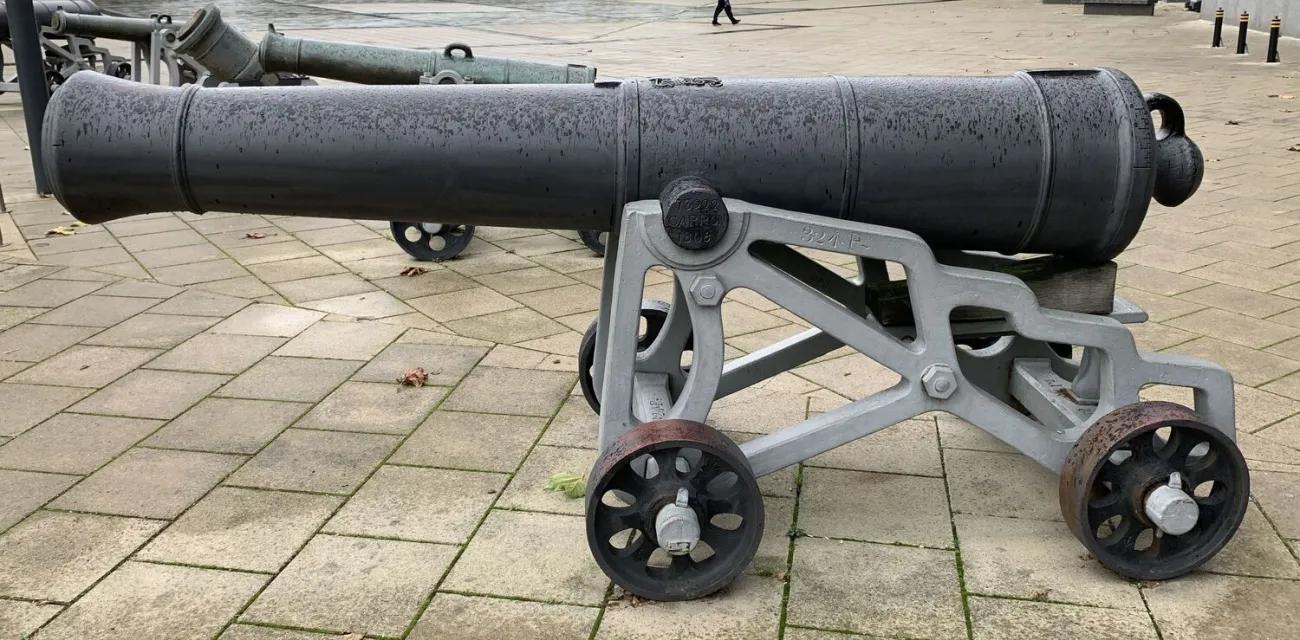 A heavy artillery barrel with raised bands and a rounded cascabel fixed to a modern grey mount with geometric supports and four metal wheels, set against a spacious paved plaza