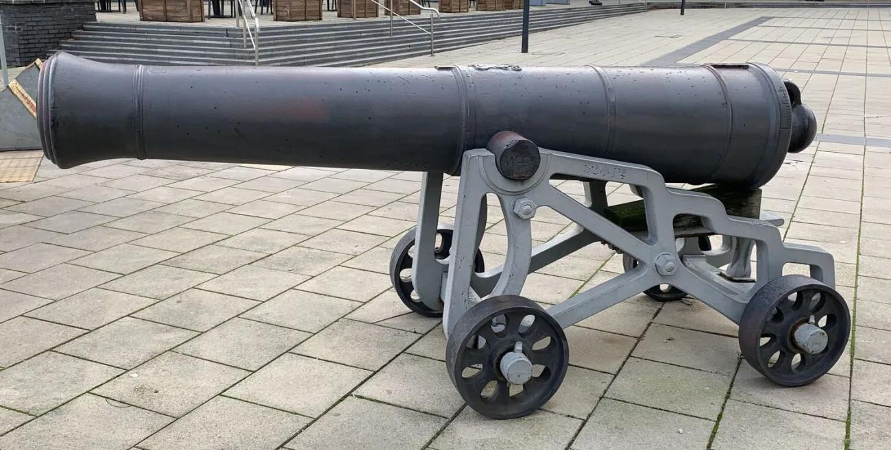 A smooth black artillery barrel resting on an angular grey metal support frame, featuring rust‑rimmed wheels and set on light square paving stones in an open public space