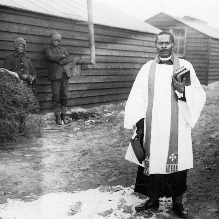 Priest in vestments is stood in a camp with wooden huts., two other soldiers are stood behind him