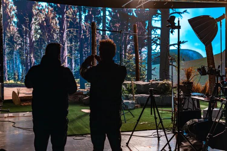 Behind the scenes of filming production with forest scene on a large screen in the background and silhouette of production team in the foreground.