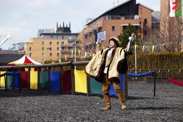 A speaker in light yellow Tudor dress delivers an announcement to the crowd in the Tiltyard