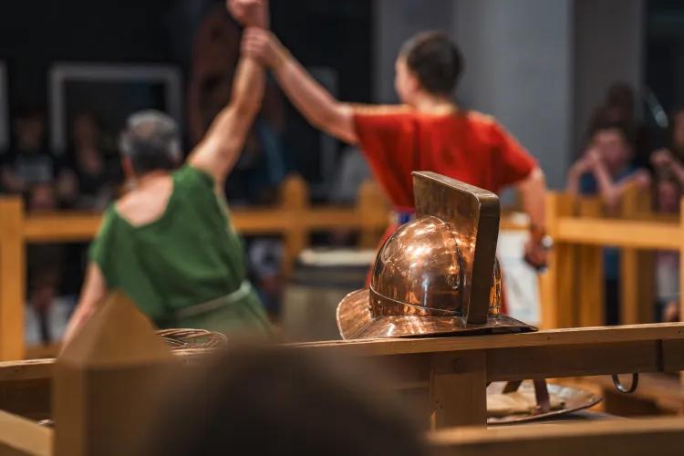 Two people in Roman tunics hold up their hands in victory. A gladiator helmet is in focus in the foreground.