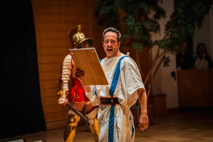 A man in a white toga with two blue stripes reads from a wooden tablet, introducing a gladiator who stands behind him