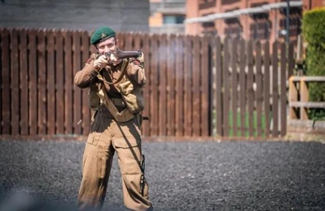 A soldier in khaki and a green beret fires a British SMLE rifle against a brown fence