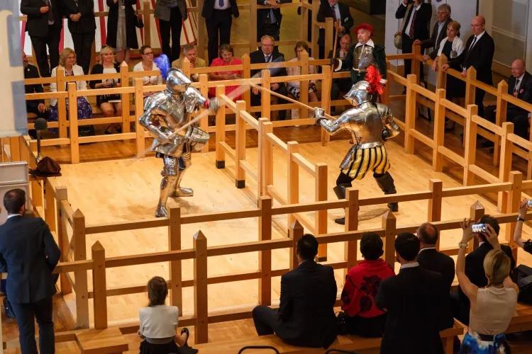 Two men dressed in half plate armour spar with each other in a wooden arena. People in suits surround them.