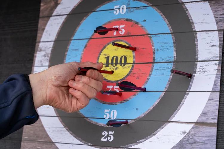 Hand holding a crossbow bolt against an archery target marked 25–100, with several bolts embedded near the centre