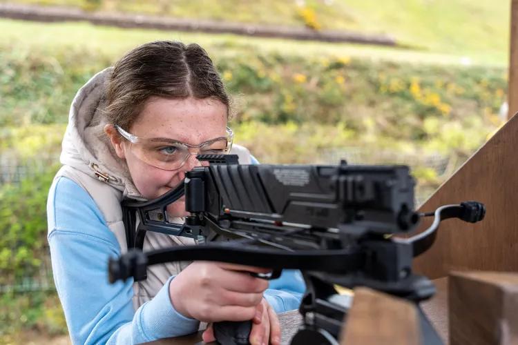 Person with face obscured aiming a mounted crossbow at a shooting range, hands gripping the stock and trigger